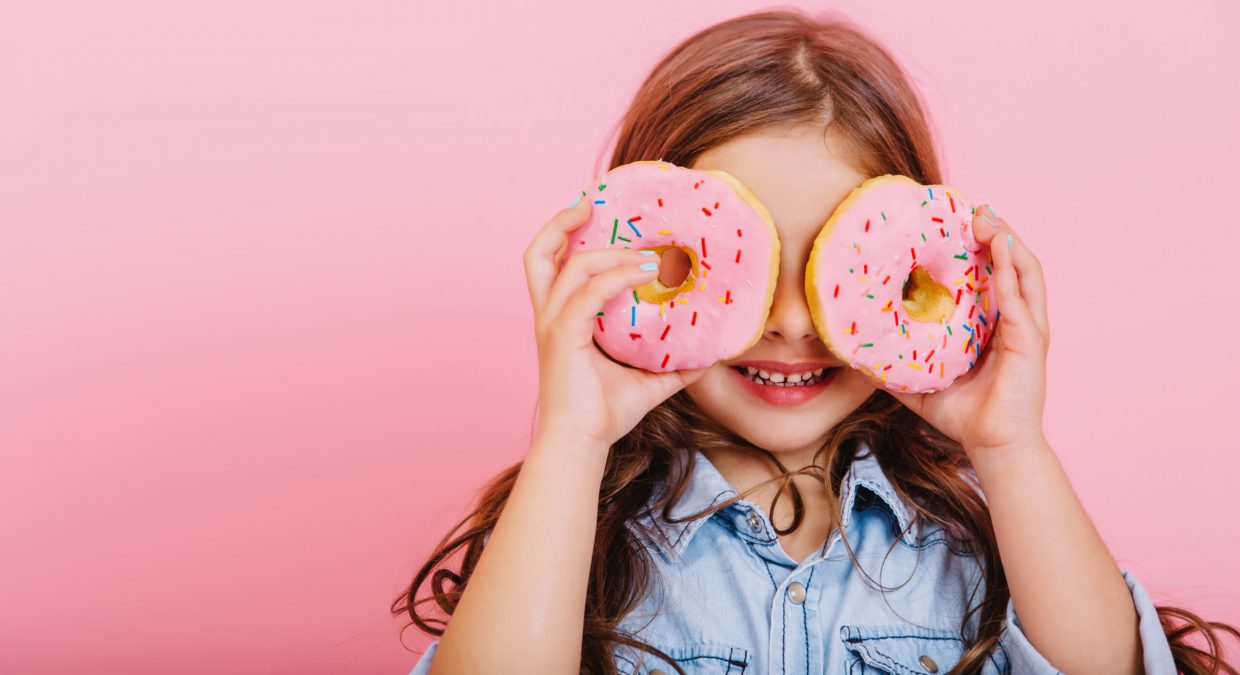 Portrait excited joyful young pretty girl in blue shirt expressing positivity, having fun to camera with donuts on eyes isolated on pink background. Happy childhood with tasty dessert. Place fot text